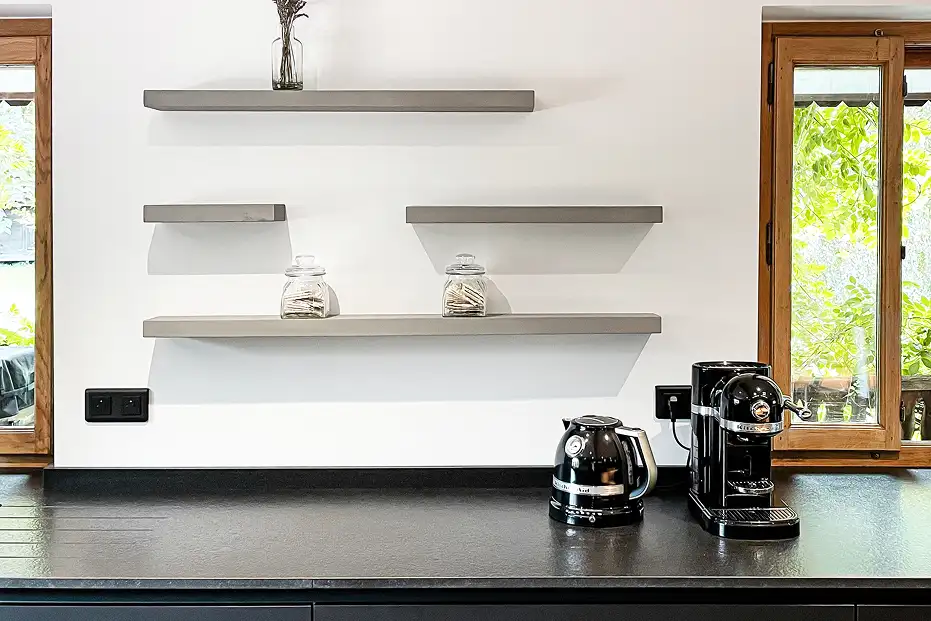 Grey concrete shelves in a kitchen between two wood windows, black countertop