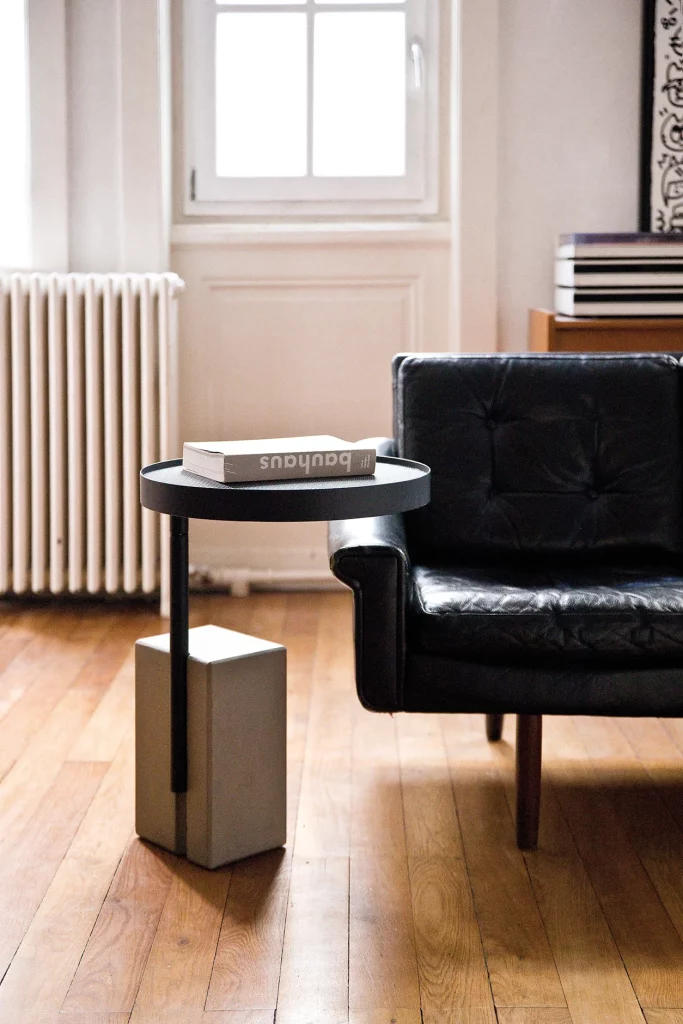 Twist side table with a black metal top on a concrete base, next to a leather armchair.